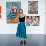 Lynda Phillips standing in front of her painting s at the 2023 Diploma Graduate's Exhibition. She's wearing a blue a-line skirt, a black top and black sandals.