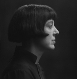 A striking black and white profile portrait of the artist, writer and illustrator Dorothy Burroughes, alumna of the Heatherley School of Fine Art. Her pale, white face and pointed nose are a stark contrast against the black background. Her black hair, in a shingle bob, it's single curl cutting into her cheek as the fringe frames her eye.